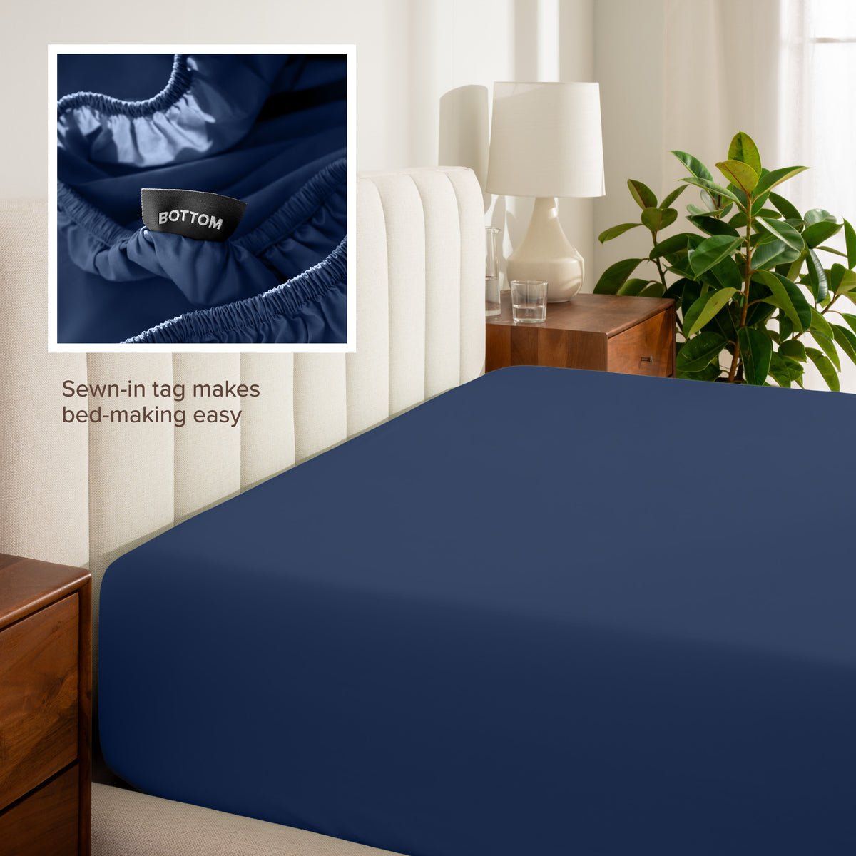 Fitted sheet on a bed with a close-up of a sewn-in tag, nightstand, lamp, and plant in a bedroom setting.