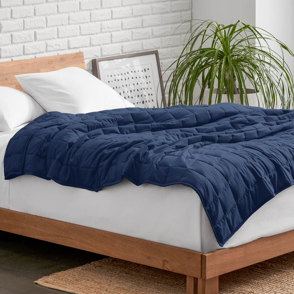 Weighted blanket on a bed with white pillows and a wooden headboard in a room with a plant and brick wall.