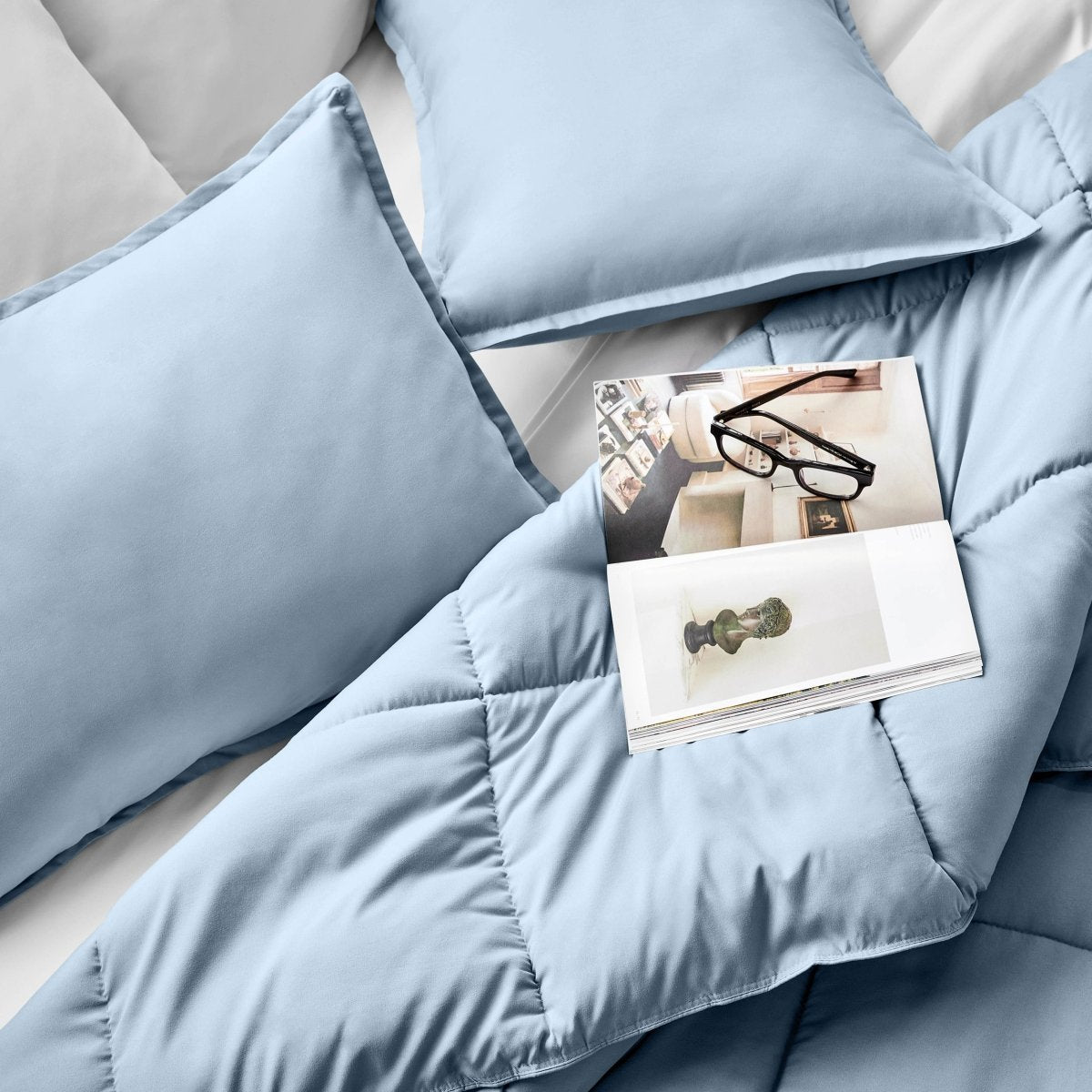 Comforter and pillows on a bed with a magazine and glasses.