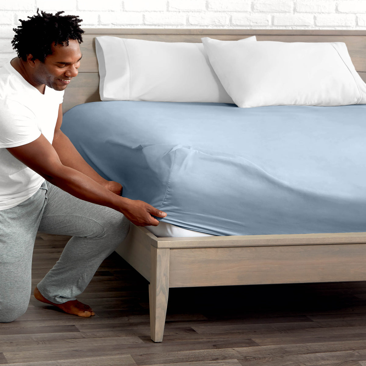 Man adjusting fitted sheet on a bed with white pillows.