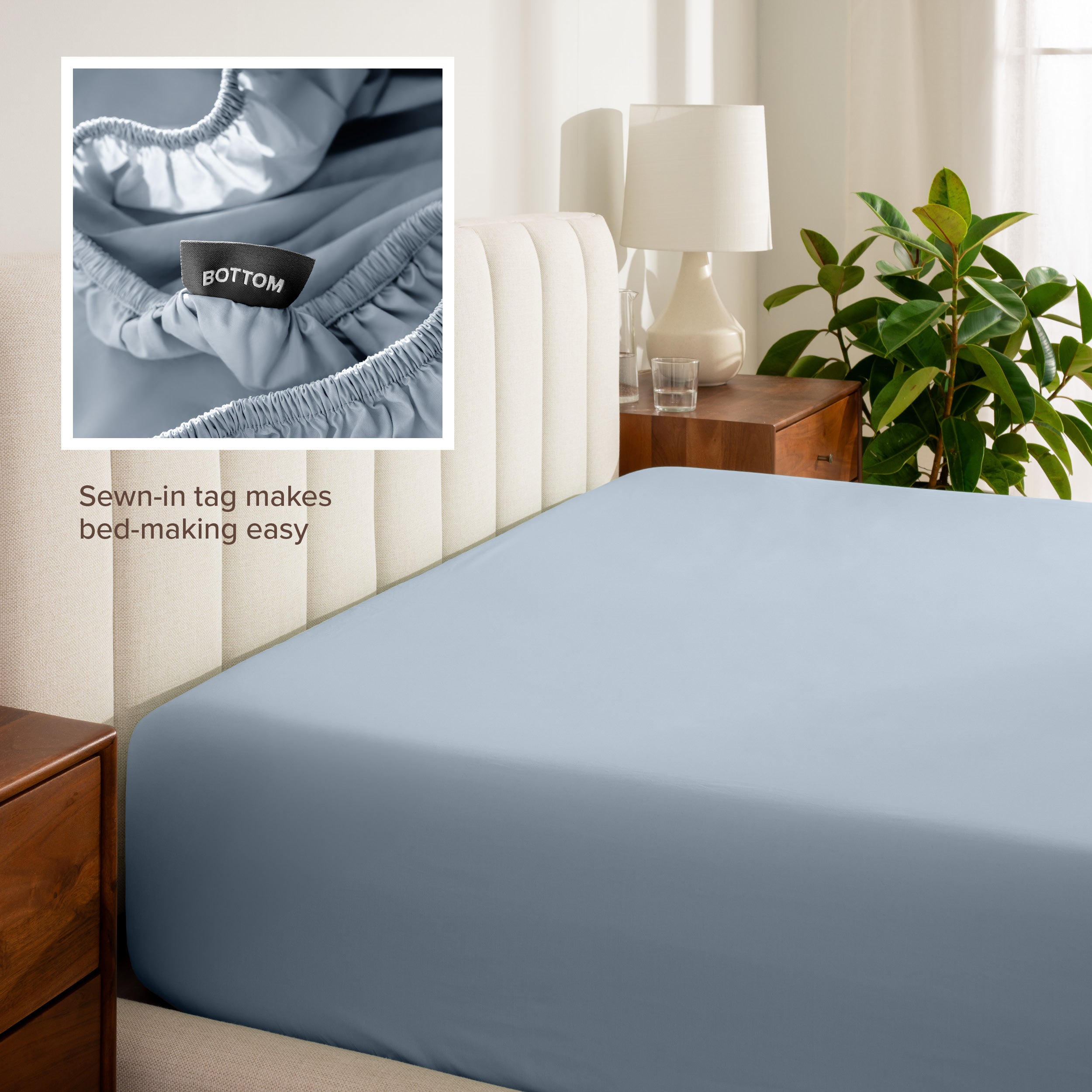 Fitted sheet on a bed with a close-up of a sewn-in tag, nightstand, lamp, and plant in a bedroom setting.