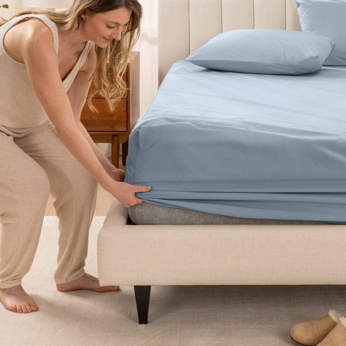 Woman in pajamas adjusting bed sheets on a bed.