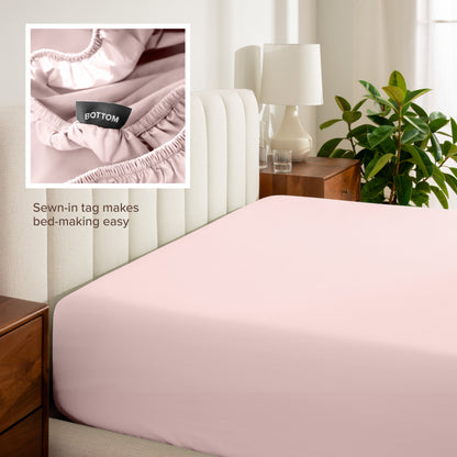 Fitted sheet on a bed with a close-up of a sewn-in tag, nightstand, lamp, and plant in a bedroom setting.