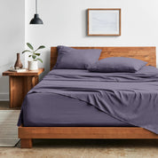 Bedroom with sheet set on a wooden bed frame, side table with plant and lamp.