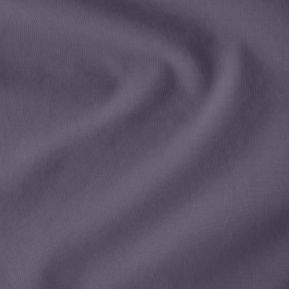 Fitted sheet on a bed with a close-up of a sewn-in tag, nightstand, lamp, and plant in a bedroom setting.