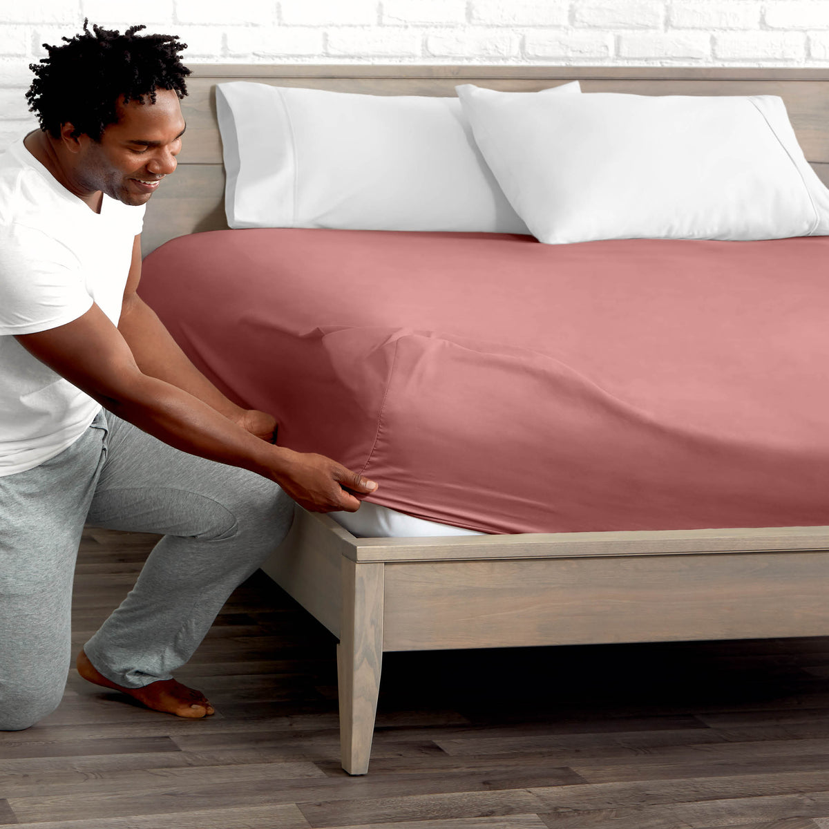 Man adjusting fitted sheet on a bed with white pillows.