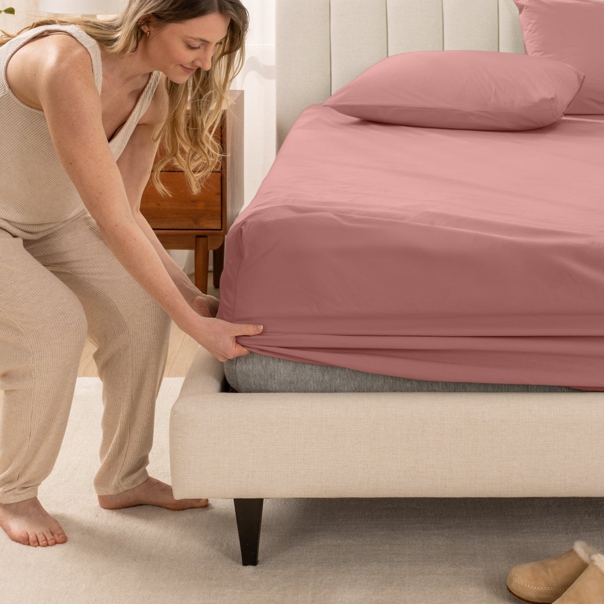 Woman in pajamas adjusting bed sheets on a bed.