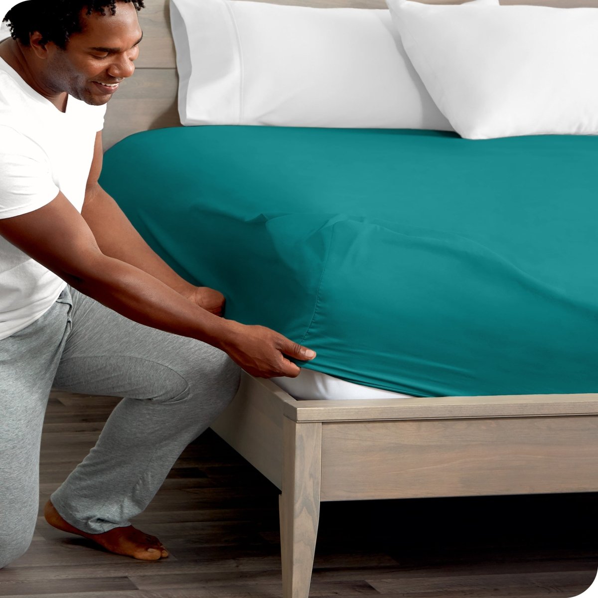 Man in white shirt and gray pants adjusting a fitted sheet on a bed.