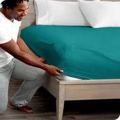 Man in white shirt and gray pants adjusting a fitted sheet on a bed.