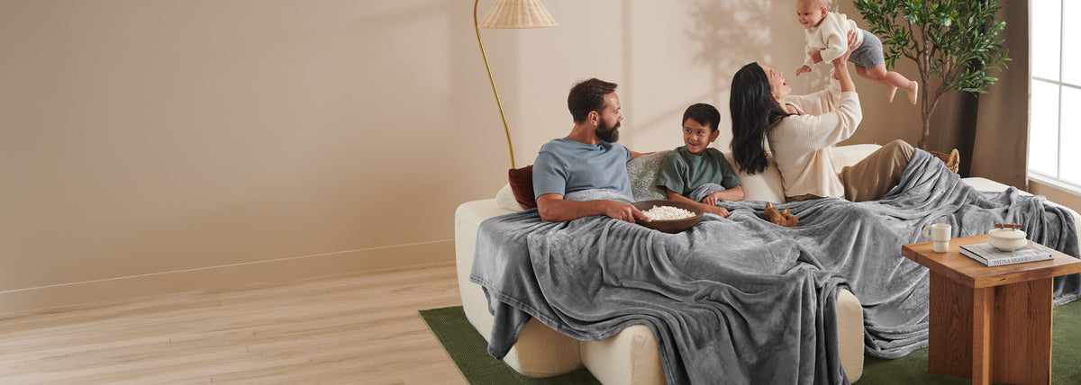 A family of four relaxes together on a cream sofa draped with a soft gray blanket in a warm, modern living room. A man on the sofa reads a book, a woman holds a baby, and another child is being lifted up by an adult in the background near a window.