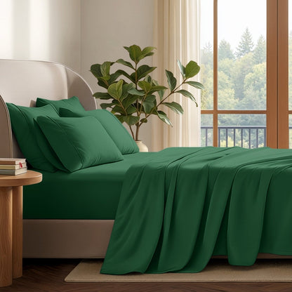 A neatly made bed with in a softly lit bedroom. A large green plant stands beside the bed, and a window with curtains is visible in the background.