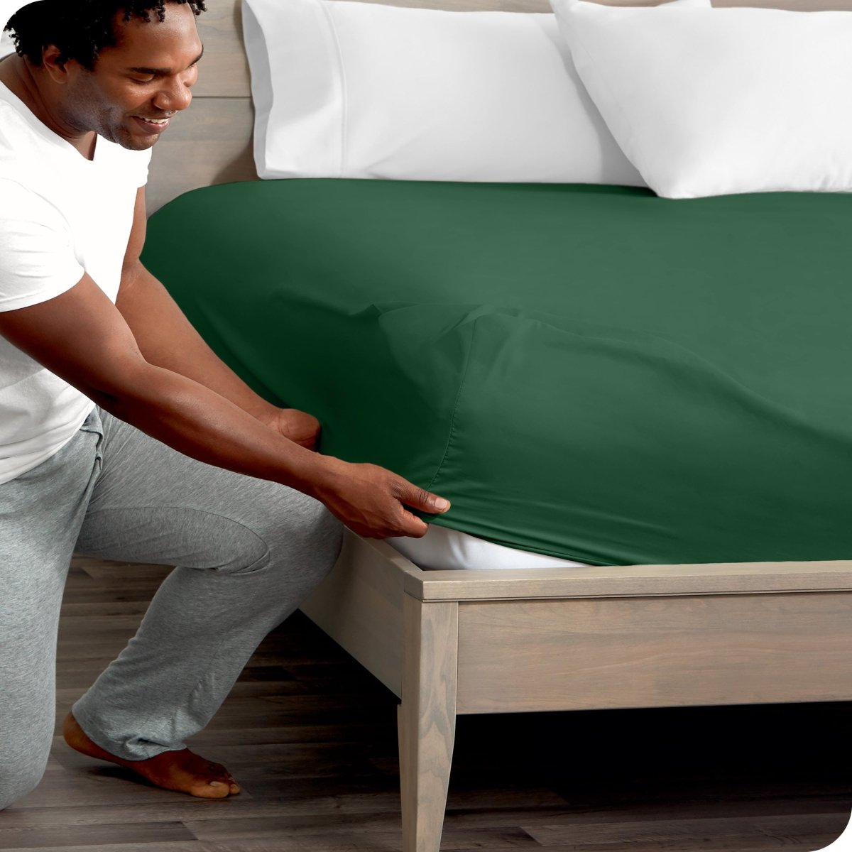 Man in white shirt and gray pants adjusting a fitted sheet on a bed.