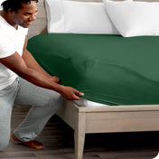 A man is kneeling while putting a fitted sheet on a bed