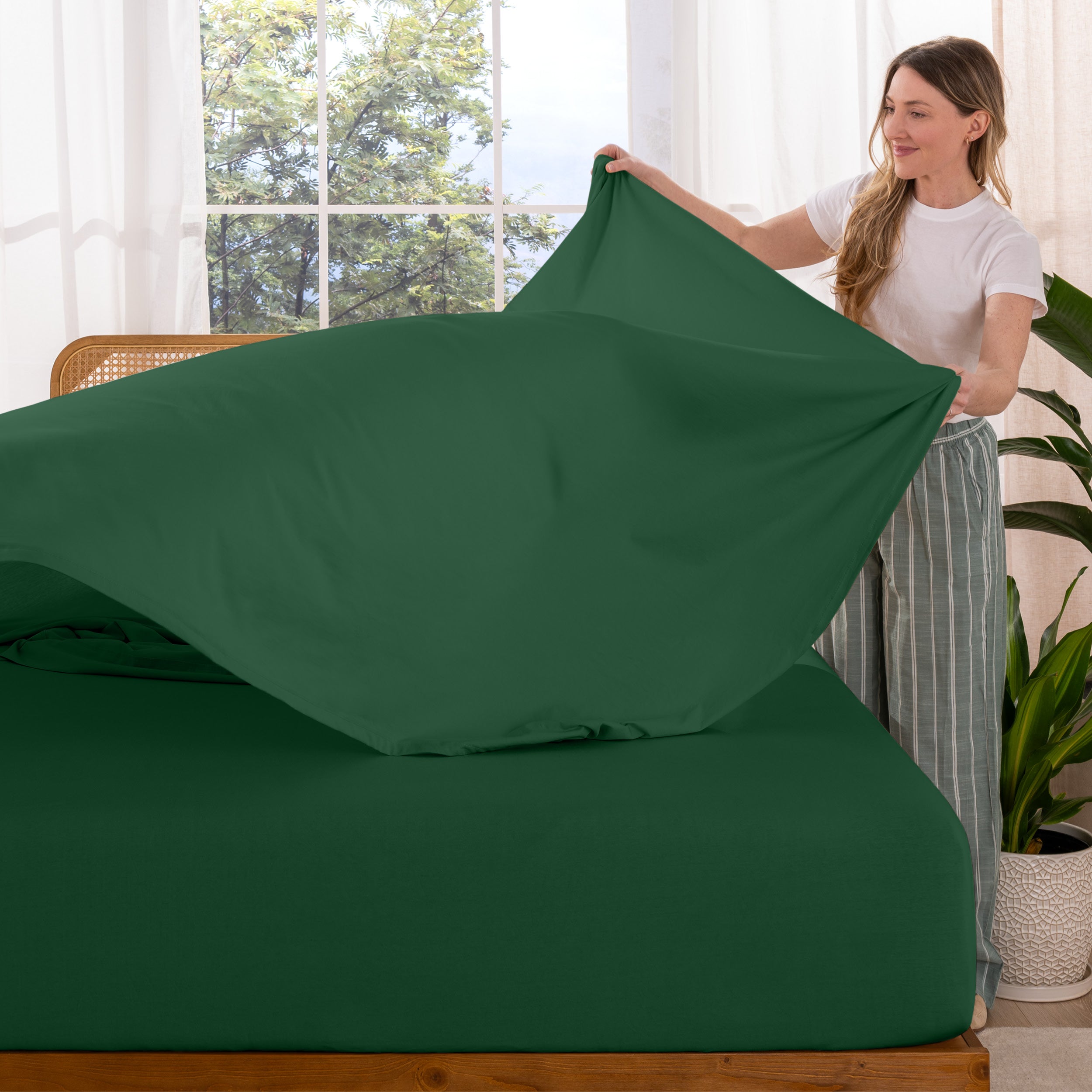 Woman putting sheet on a bed in a bright room with large windows.