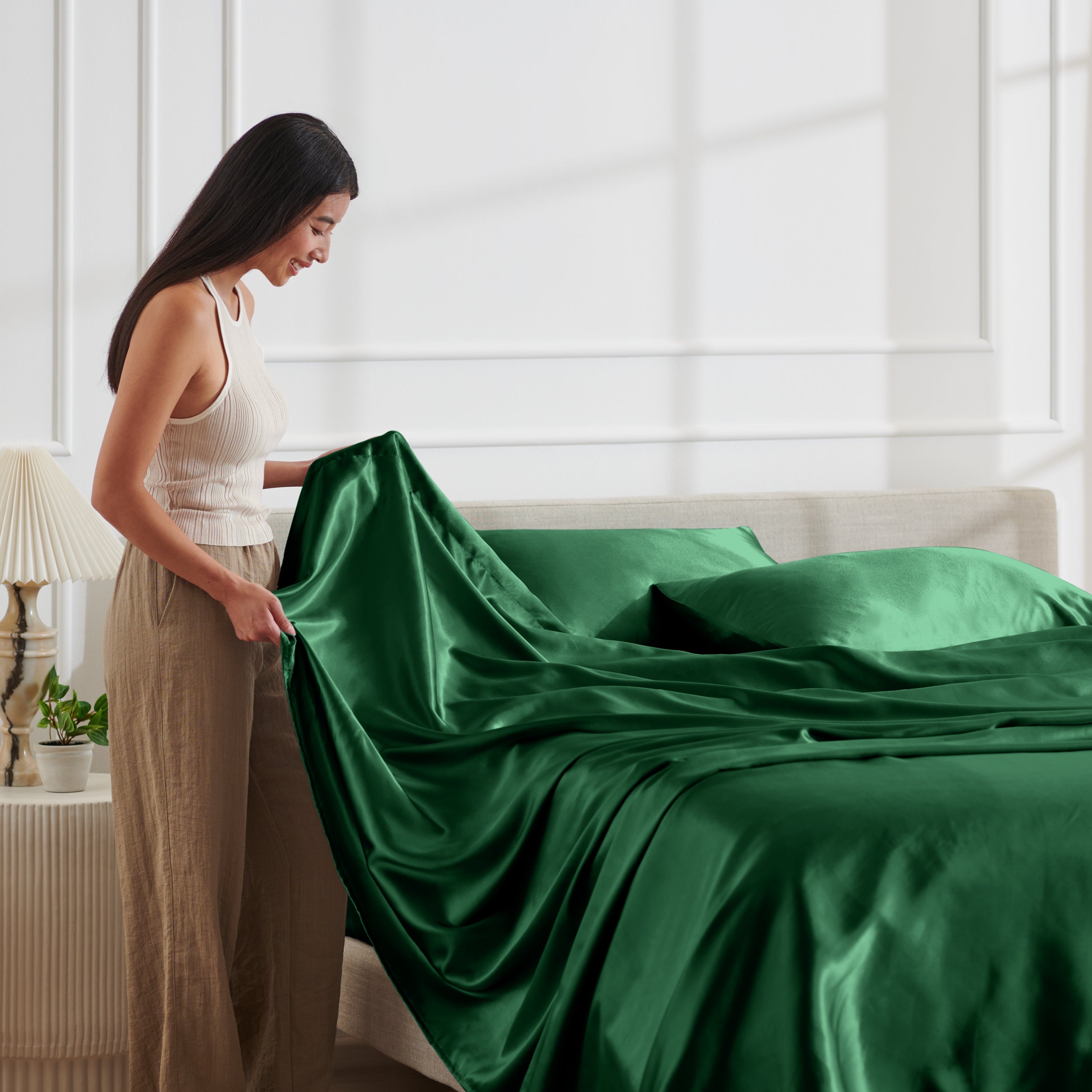 Woman arranging satin bedding on a bed in a bright bedroom.