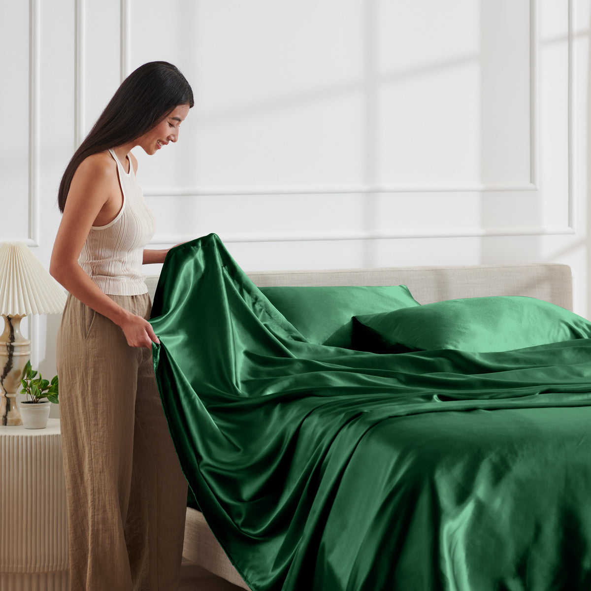 Woman arranging satin bedding on a bed in a bright bedroom.