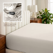 Fitted sheet on a bed with a close-up of a sewn-in tag, nightstand, lamp, and plant in a bedroom setting.