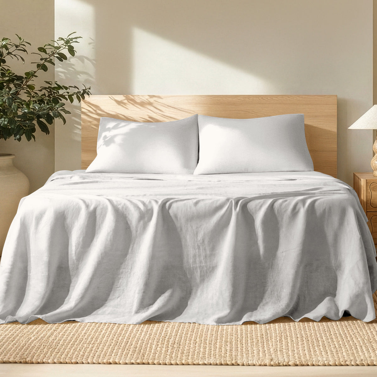 Bed with linen bedding and pillows in a bright room with a plant and lamp.