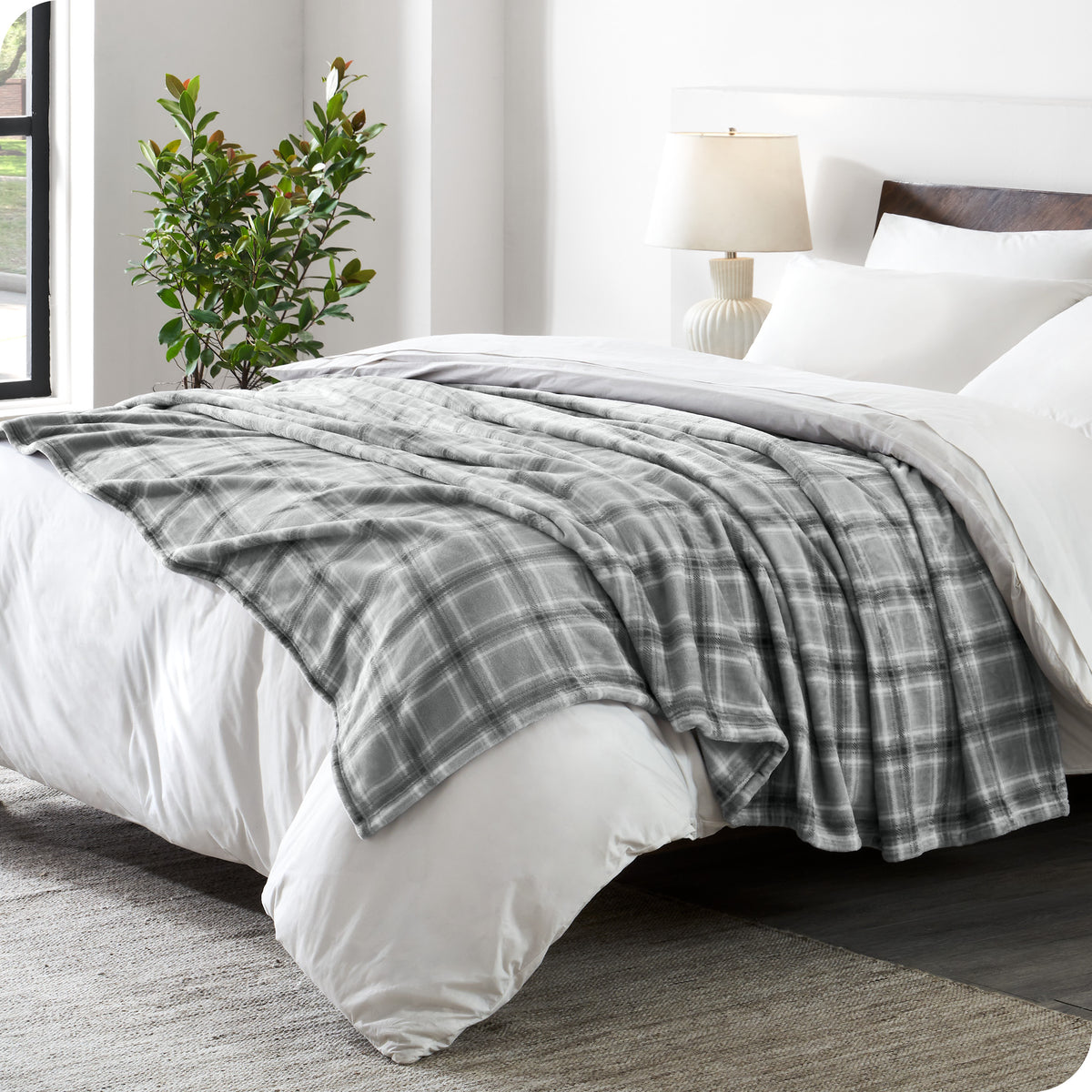 Gray plaid blanket on a white bed in a bedroom setting with a lamp and plant.
