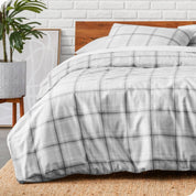 Gray plaid duvet cover on a bed with a wooden headboard and white brick wall in the background.