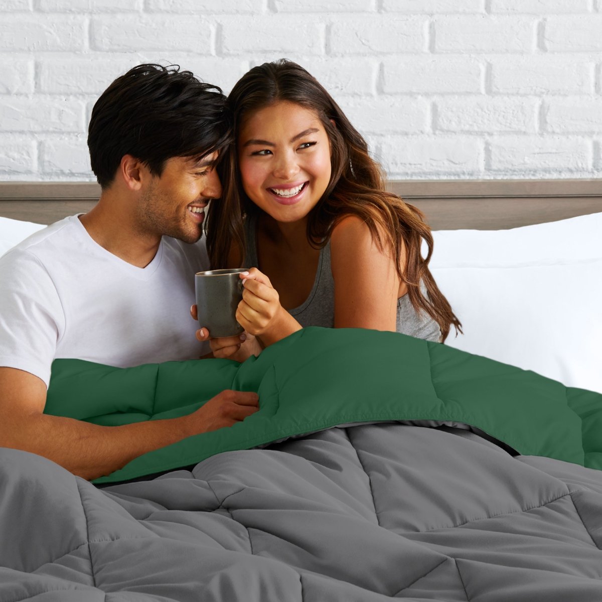 Man and woman sitting on a bed holding a mug, smiling at each other.