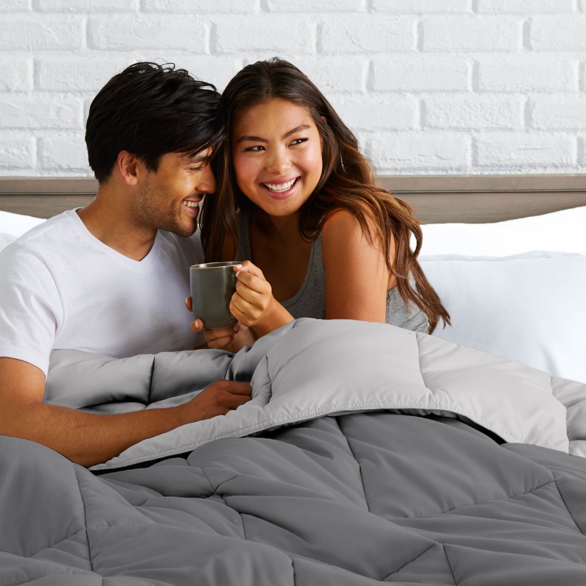 Man and woman sitting on a bed holding a mug, smiling at each other.