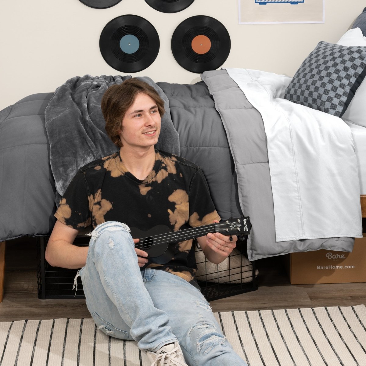 Person sitting on a bed holding a guitar in a bedroom setting with vinyl records on the wall.