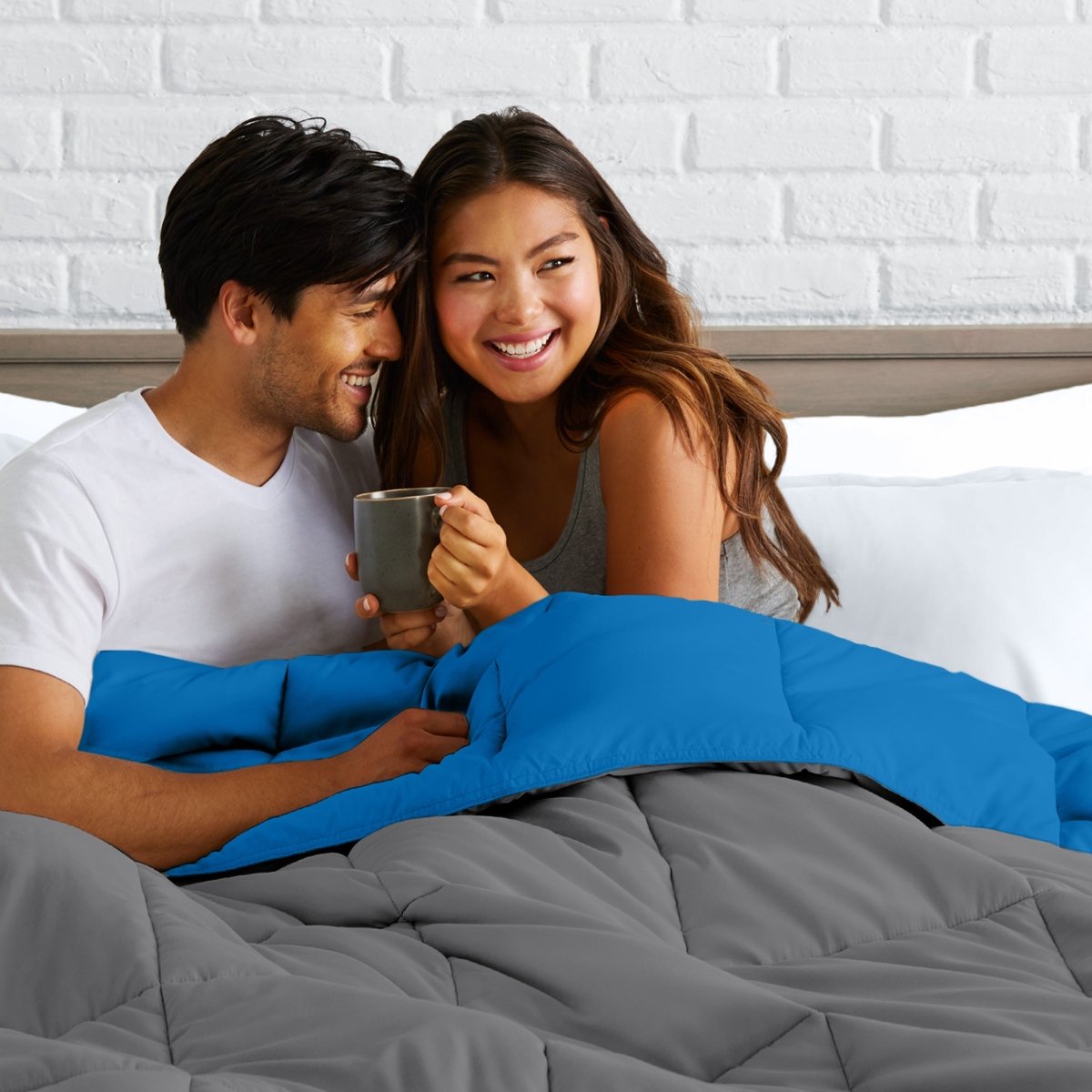 Man and woman sitting on a bed holding a mug, smiling at each other.