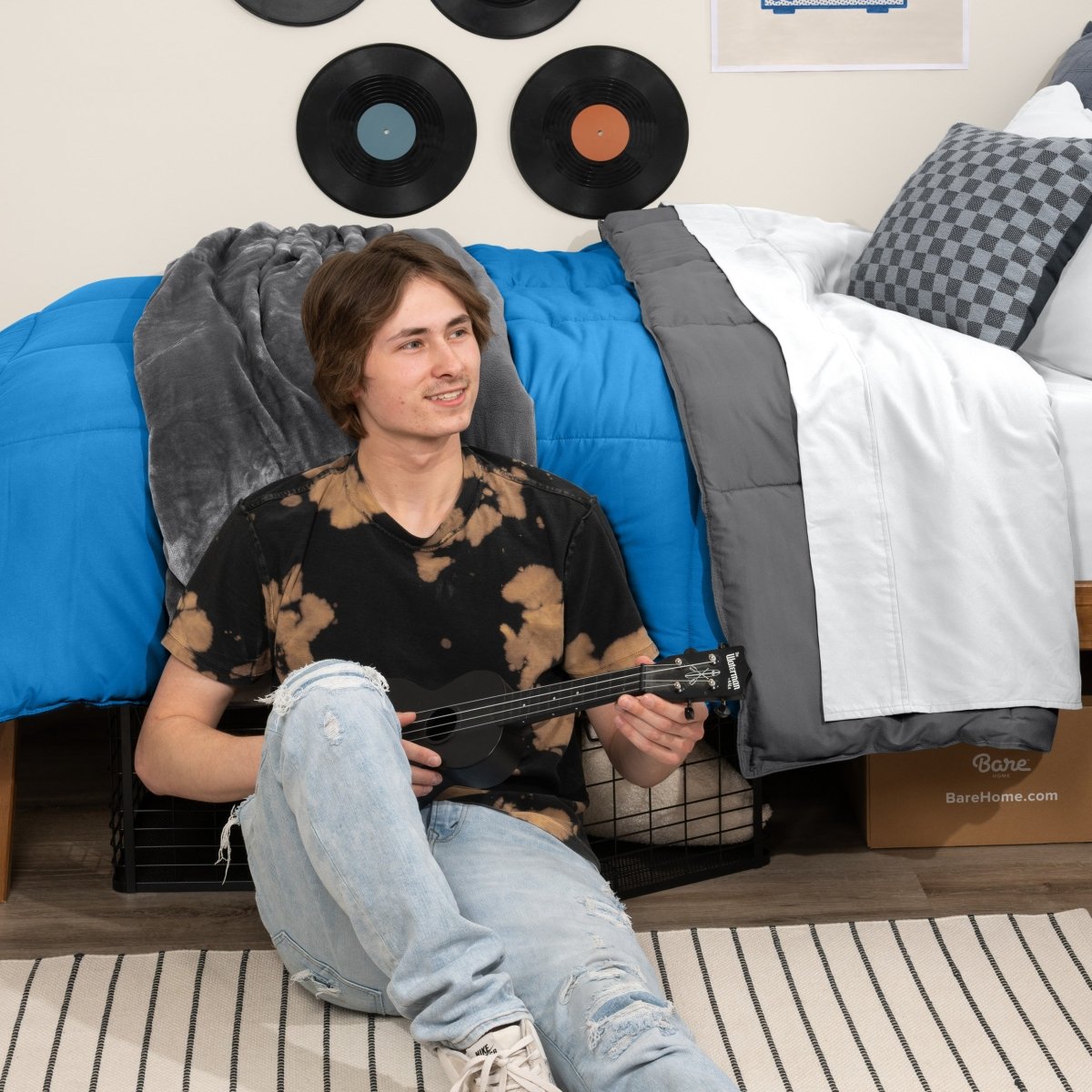 Person sitting on a bed holding a guitar in a bedroom setting with vinyl records on the wall.
