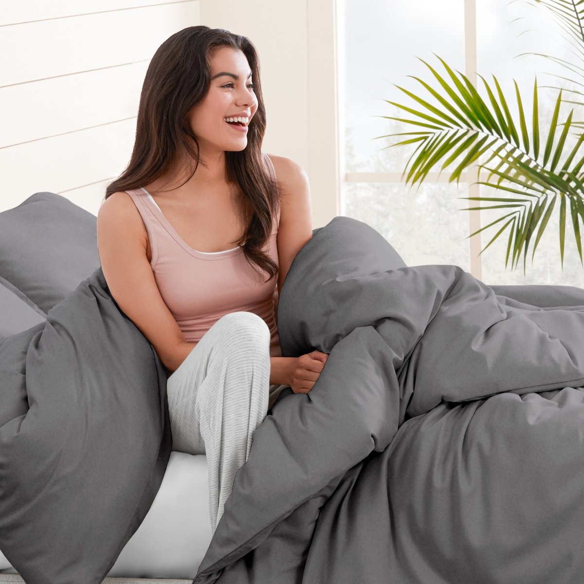 Woman sitting on a bed under duvet cover with a plant in the background