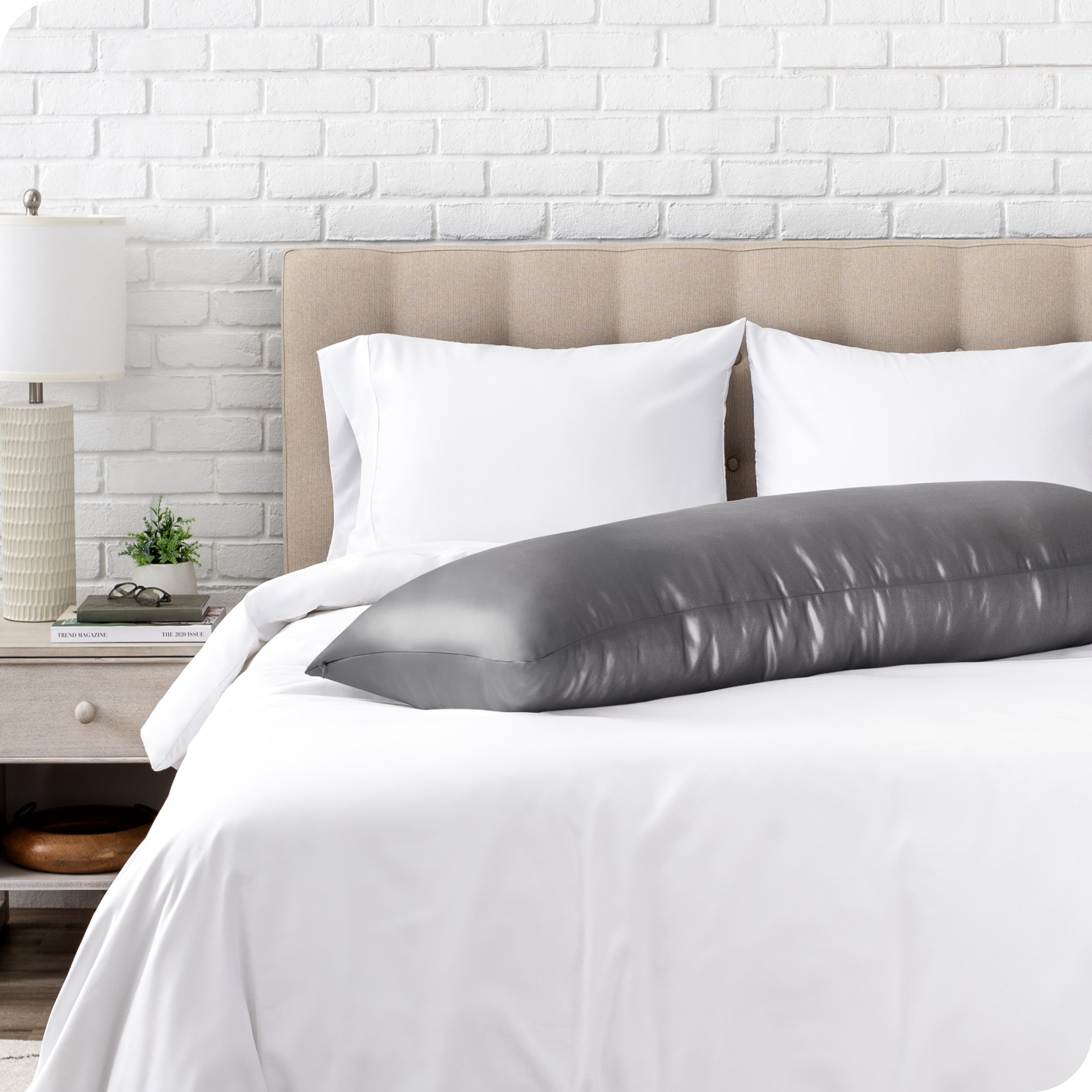 Bed with white bedding and a body pillow against a white brick wall.