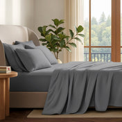 A neatly made bed with in a softly lit bedroom. A large green plant stands beside the bed, and a window with curtains is visible in the background.