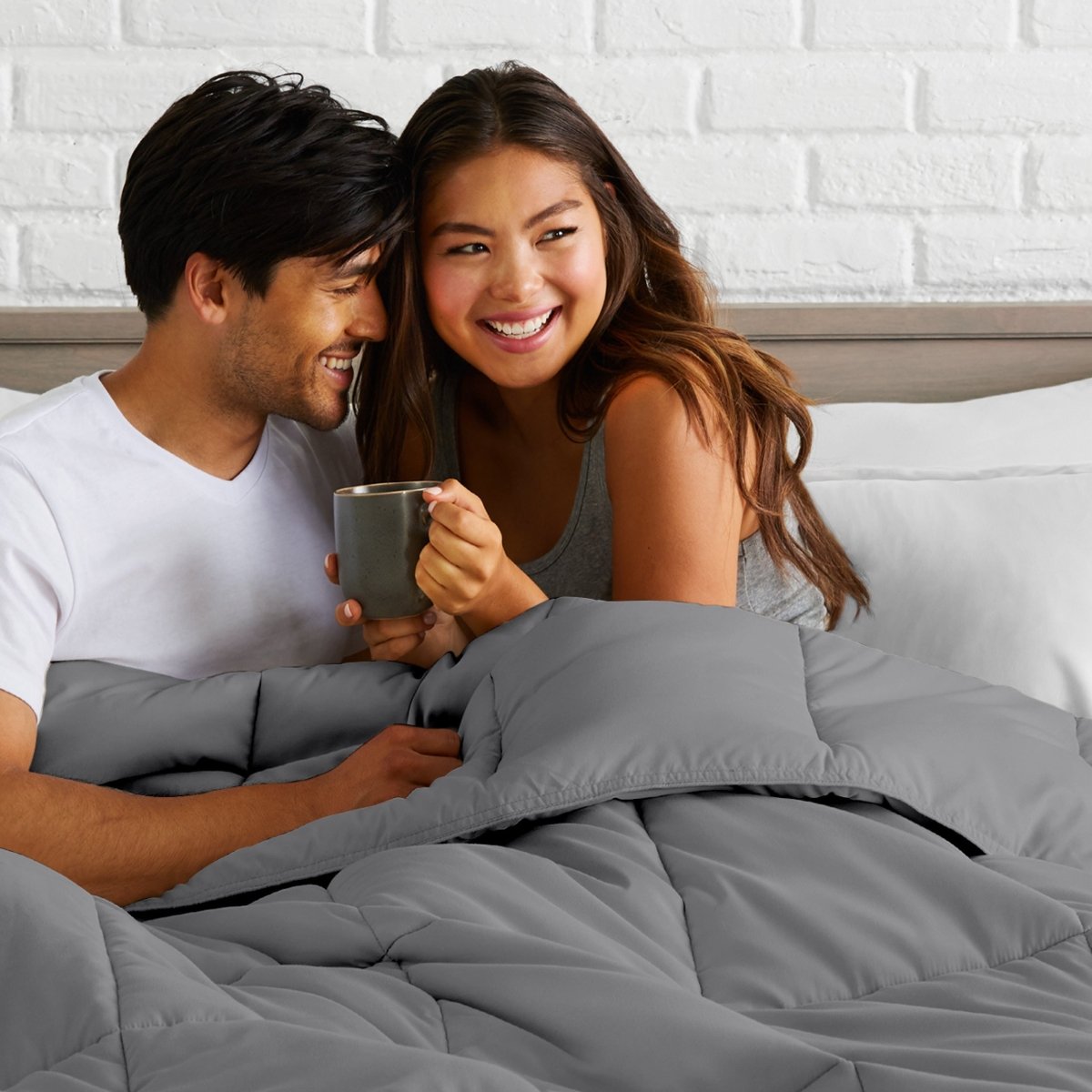 Man and woman sitting together in bed holding a mug, smiling.