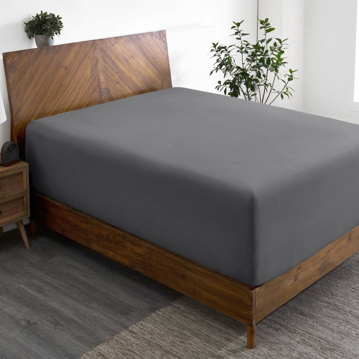 Wooden bed frame with a fitted sheet on the mattress in a room with plants and a nightstand.