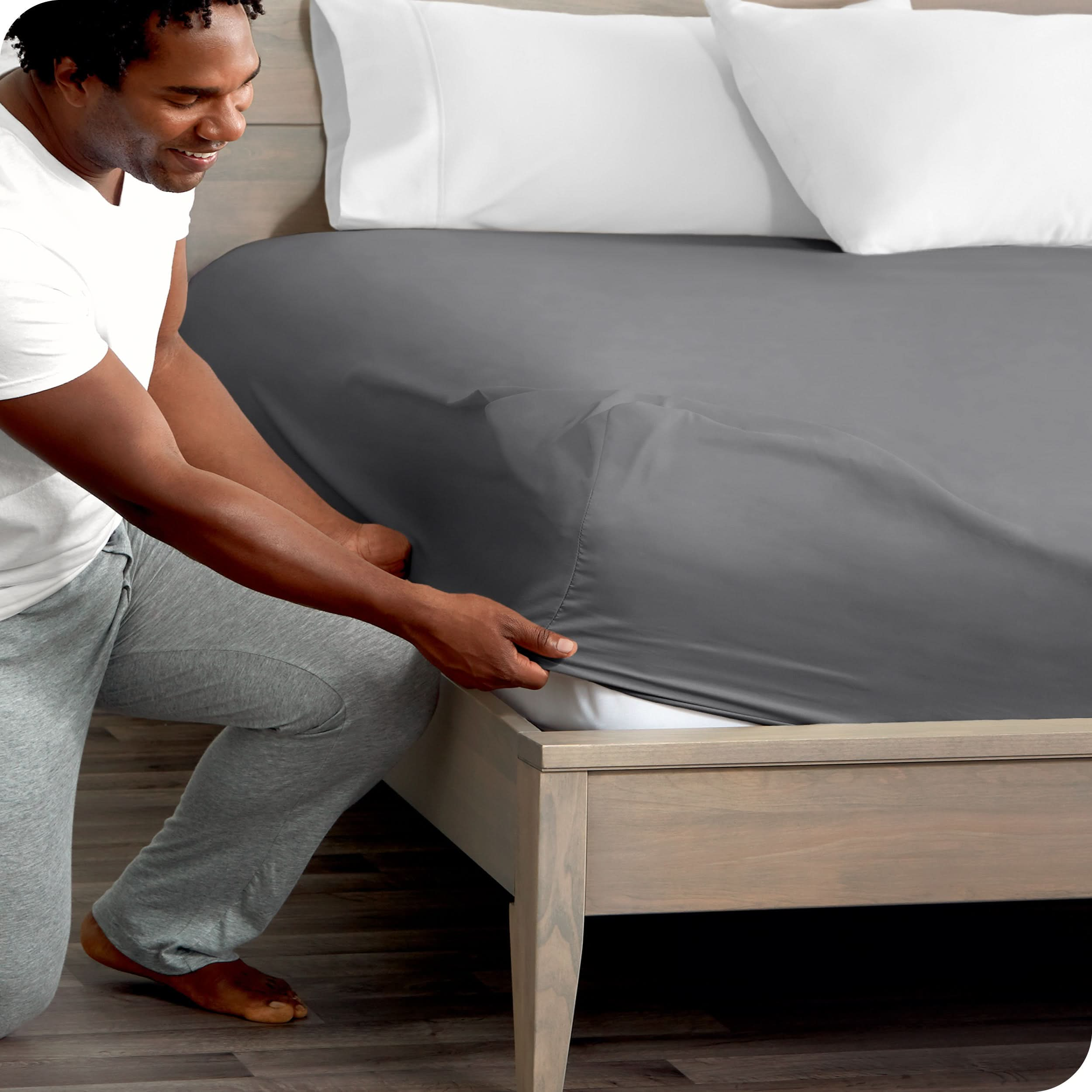 A man is kneeling while putting a fitted sheet on a bed