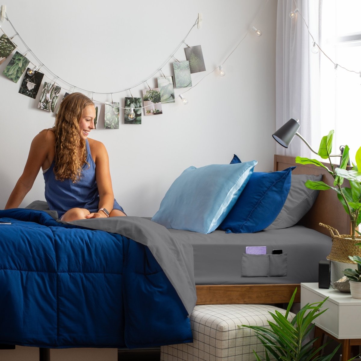 Woman sitting on a bed with blue and gray bedding in a room with plants and string lights.
