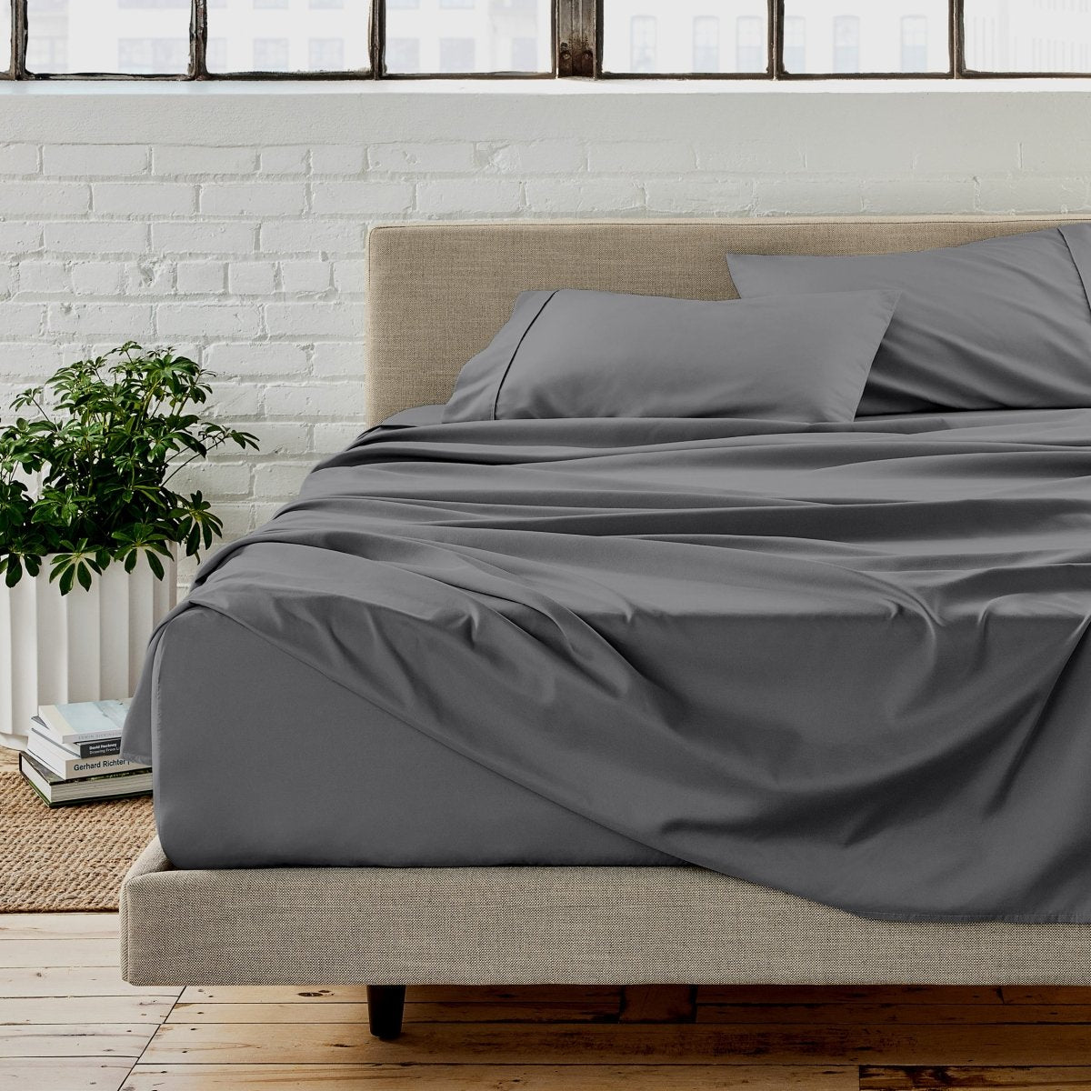 Gray sheet set on a bed in a room with a white brick wall and plants.