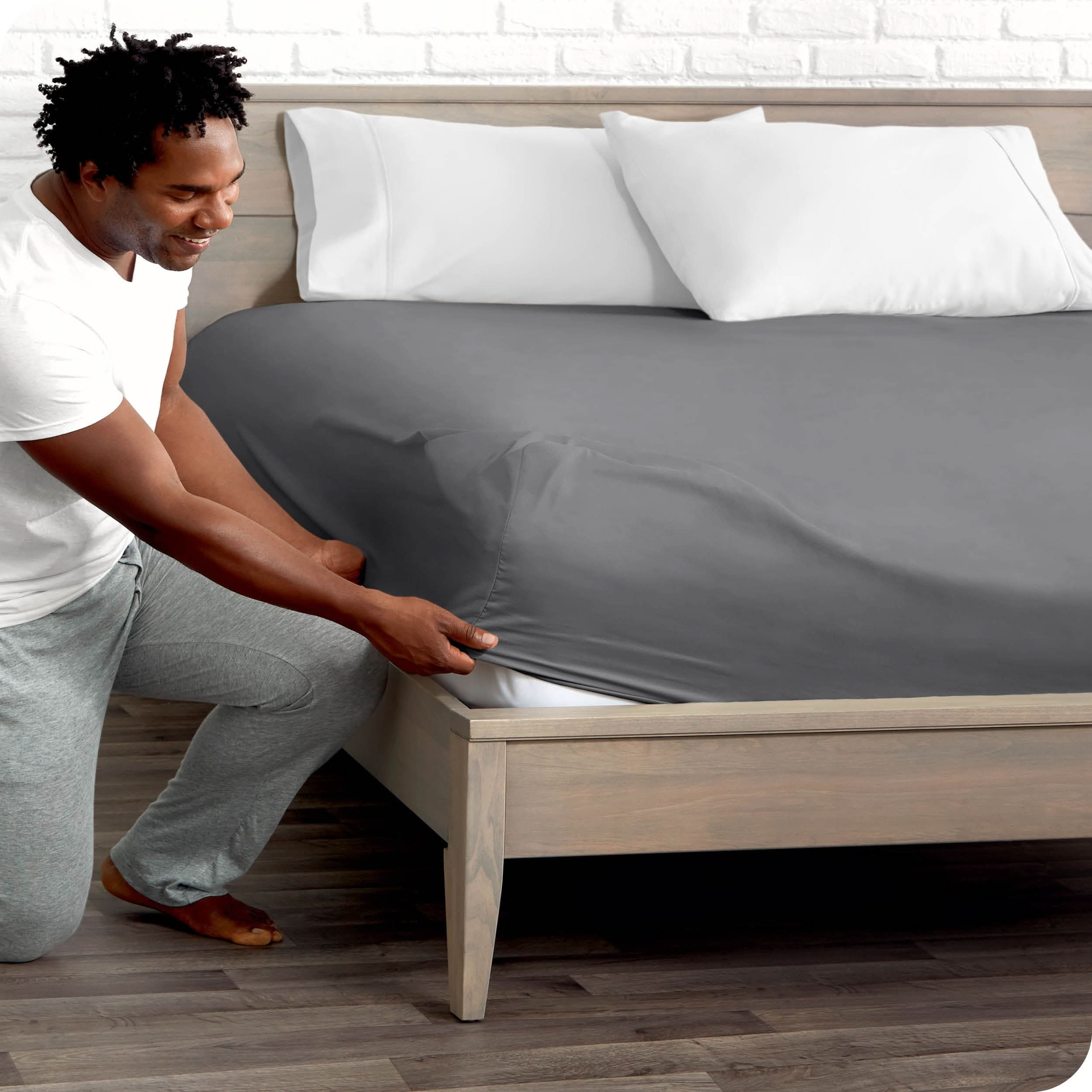 A man is kneeing and putting the corner of the fitted sheet on the mattress. The mattress is on a wooden bed frame.