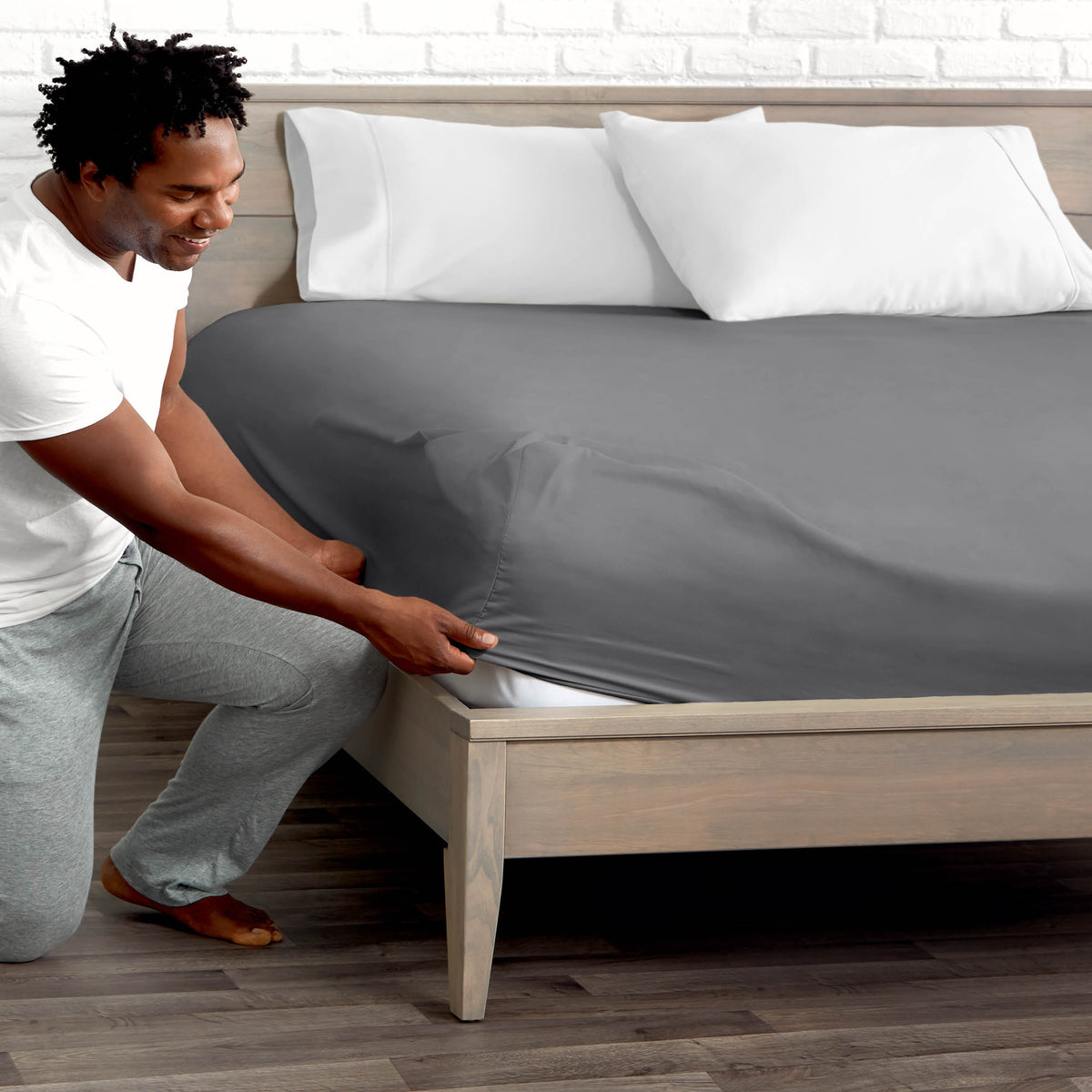 Man adjusting fitted sheet on a bed with white pillows.