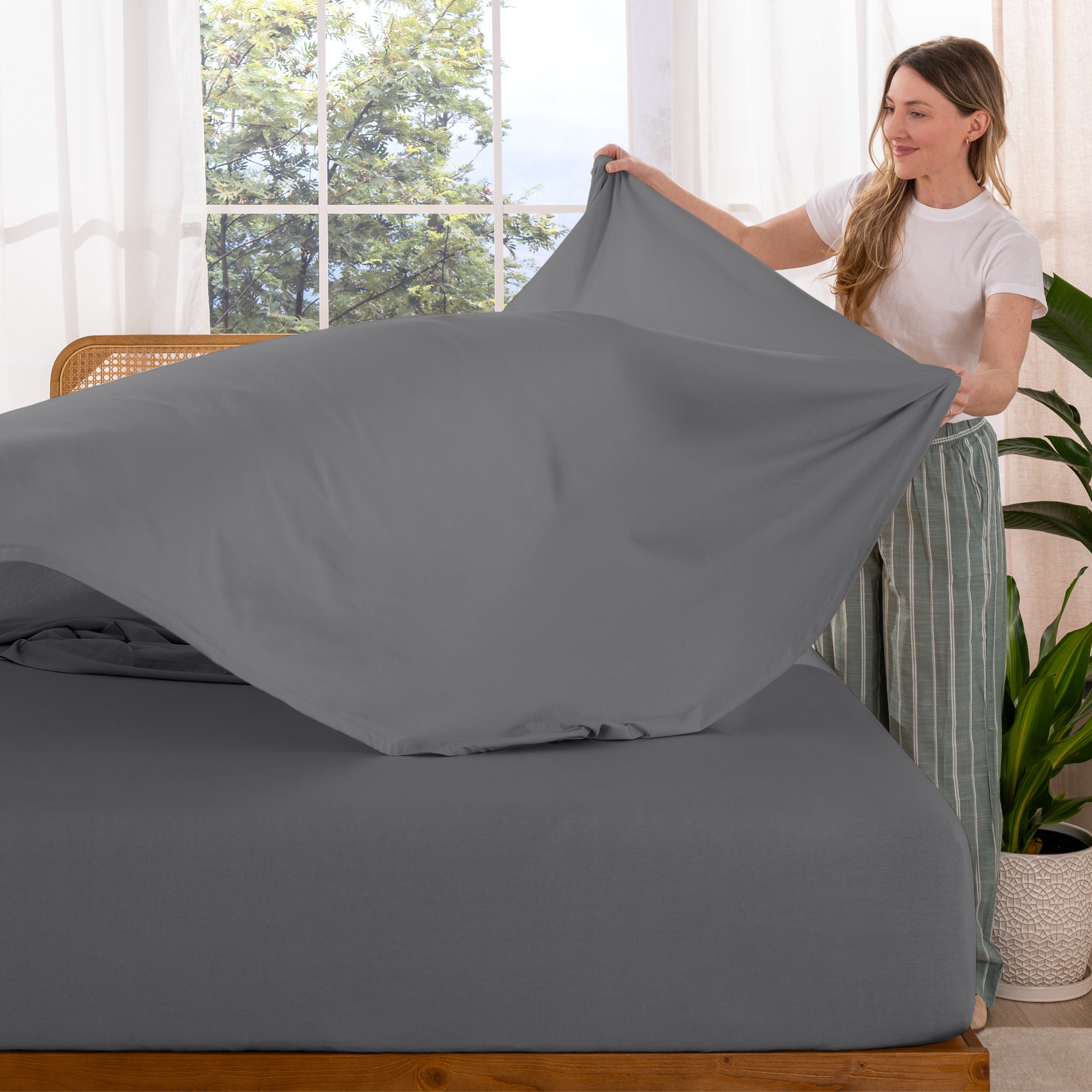 Woman putting sheet on a bed in a bright room with large windows.