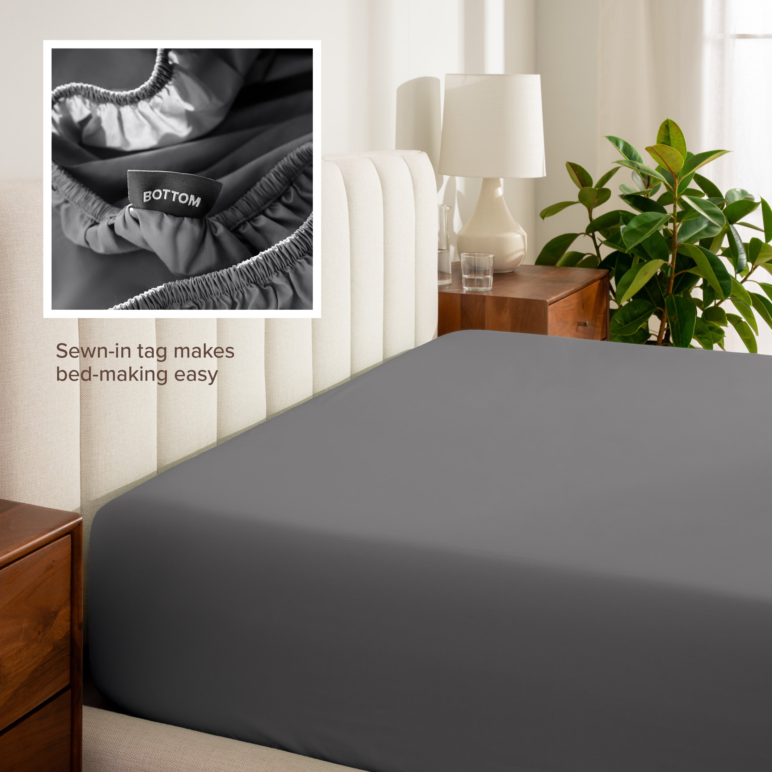 Fitted sheet on a bed with a close-up of a sewn-in tag, nightstand, lamp, and plant in a bedroom setting.