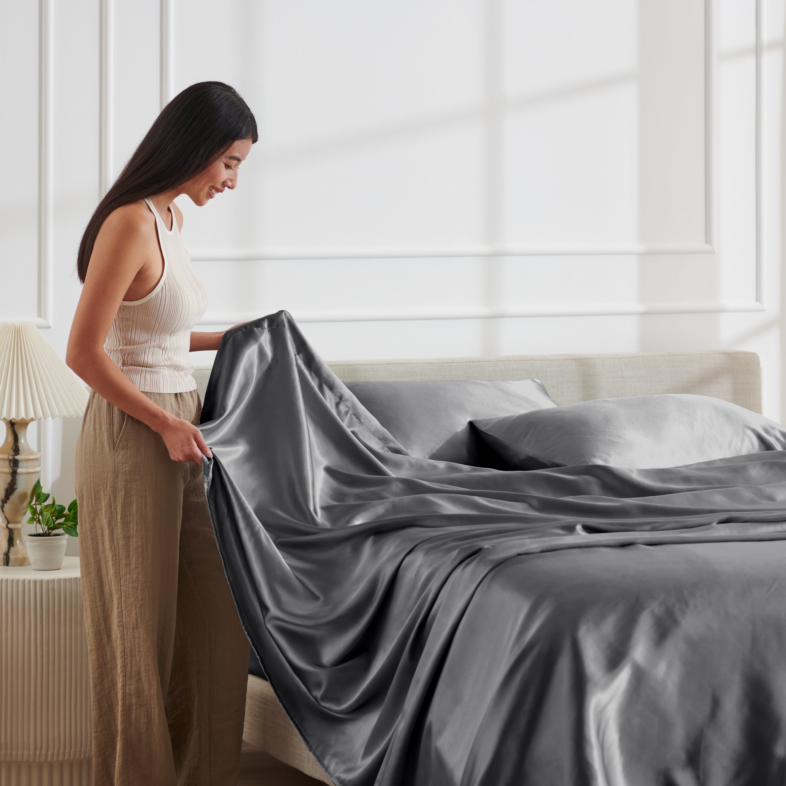 Woman arranging satin bedding on a bed in a bright bedroom.