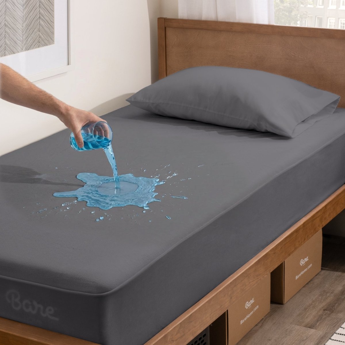 Person pouring liquid onto a gray mattress protector in a bedroom setting.
