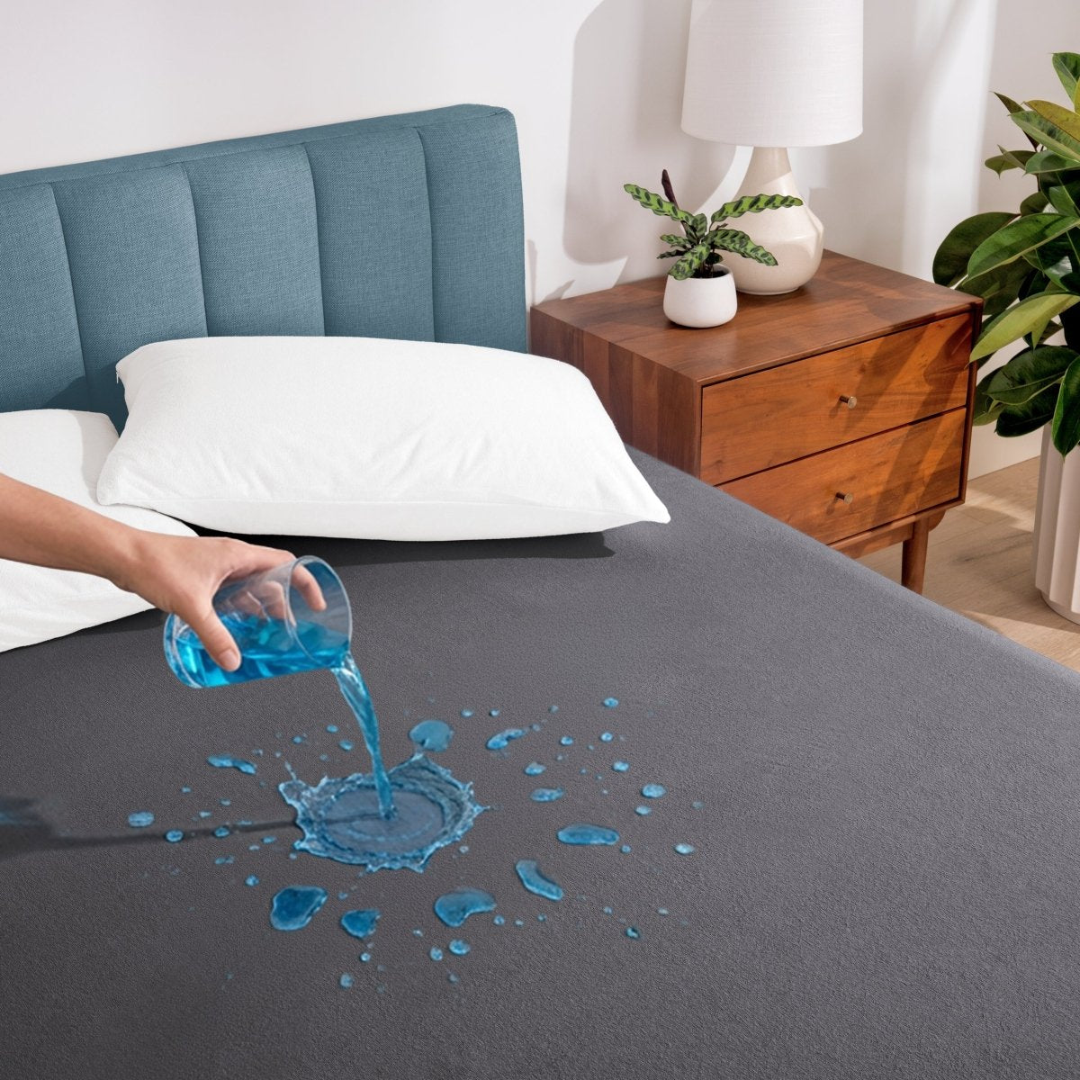 Person pouring liquid onto a gray mattress protector in a bedroom setting.