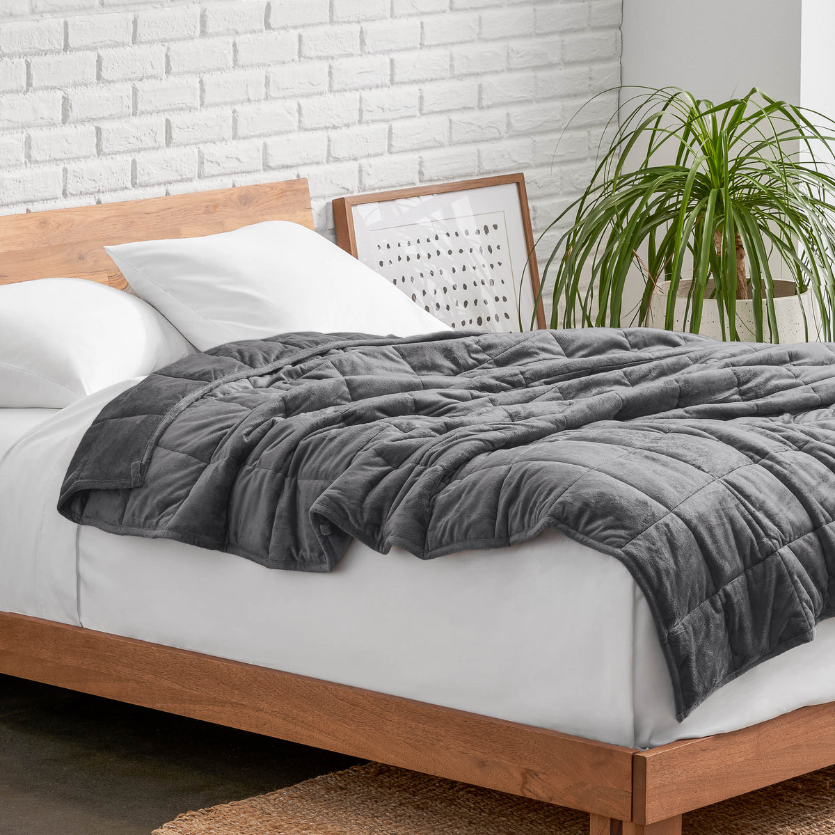 Weighted blanket on a bed with white pillows and a wooden headboard in a room with a plant and brick wall