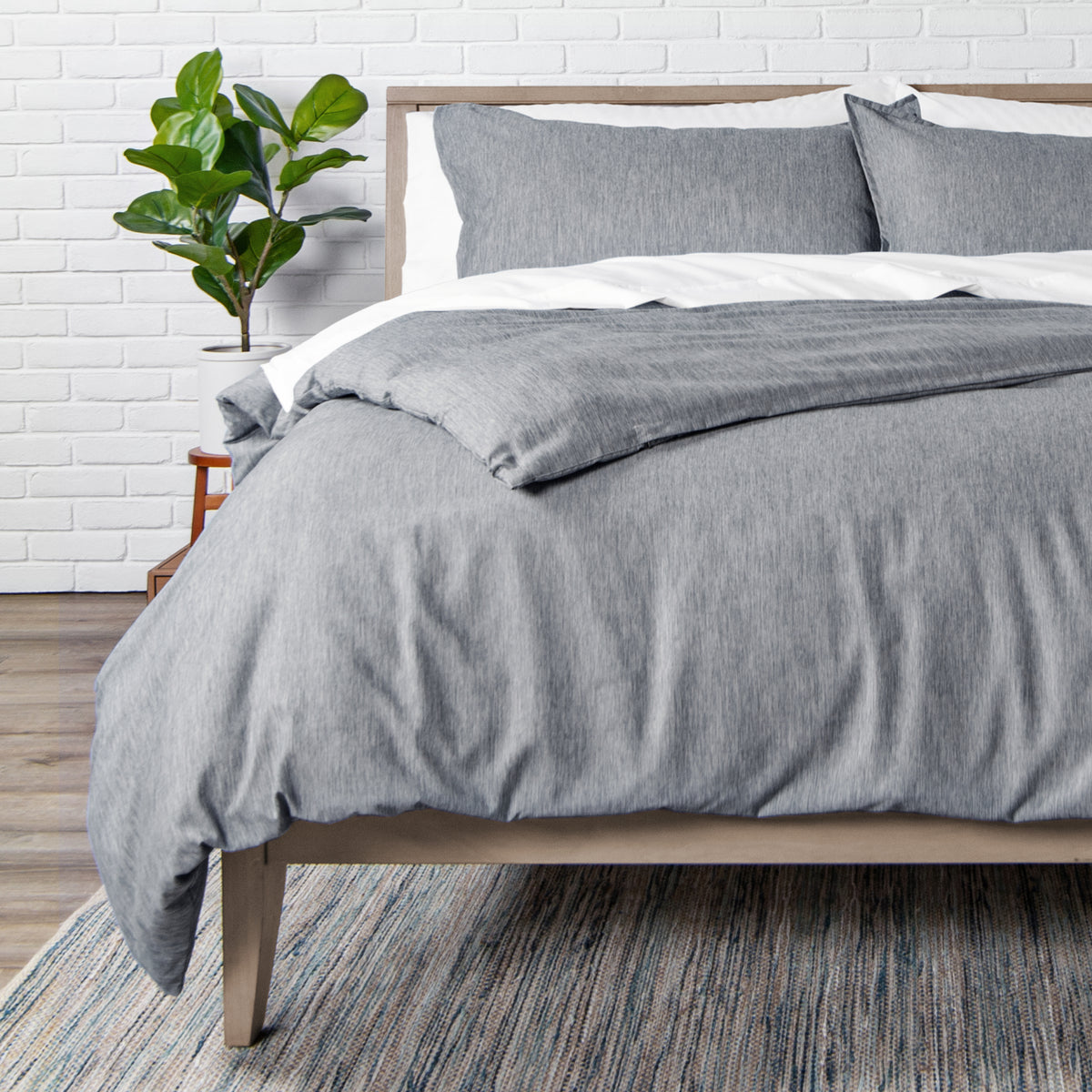 Bed with heathered charcoal bedding in a room with a white brick wall and a plant.
