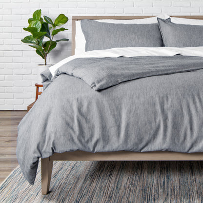 Bed with heathered charcoal bedding in a room with a white brick wall and a plant.