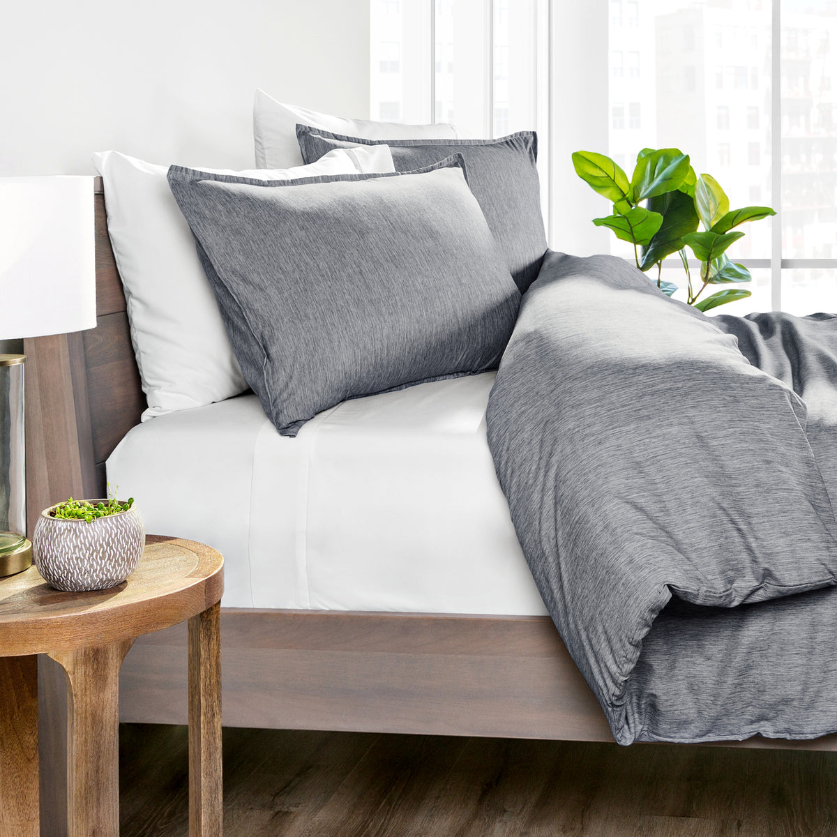 Gray bedding set on a bed with a wooden side table and plant in the background