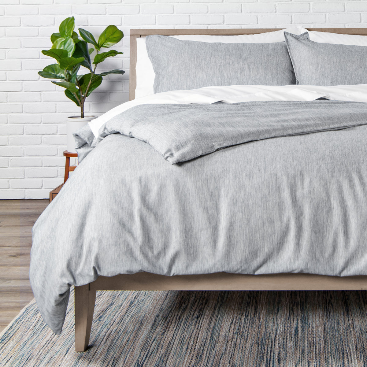 Bed with gray bedding in a room with a white brick wall and a plant.