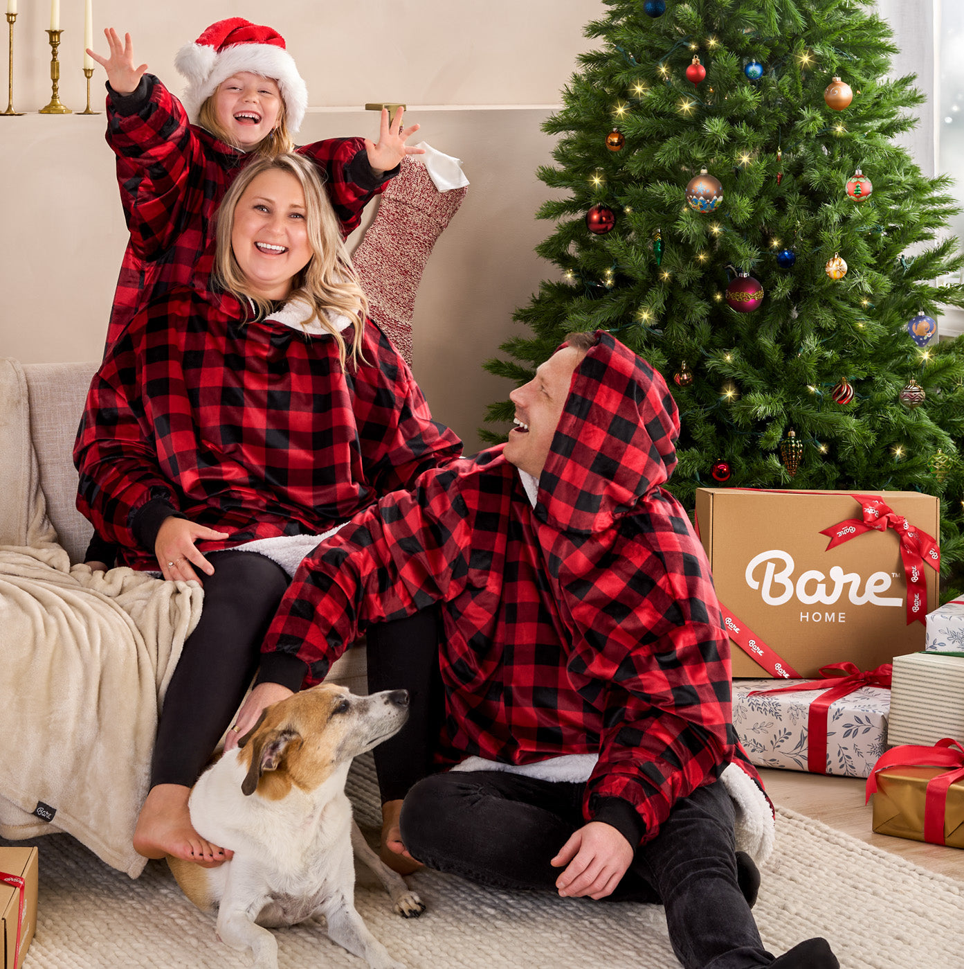 Family in red and black plaid blanket hoodies sitting on the floor in front of a decorated Christmas tree with Bare Home branding.